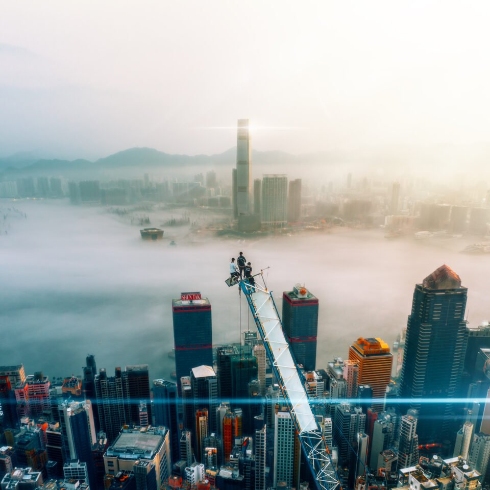 Three adventurous climbers stand triumphantly at the top of a towering crane in Hong Kong, with the city skyline and mist-covered ocean visible in the background. Their daring feat is captured in this stunning image, which showcases the thrill of exploration and the beauty of the urban landscape.